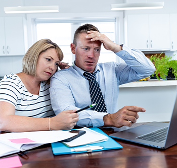 Two adults sit at a wooden table in a bright kitchen or dining area while looking at a laptop computer. Papers, folders, sticky notes, a pen, and a smartphone are spread across the table. One person points toward the laptop screen while the other leans closer to view it. White cabinets, windows, and a potted plant are visible in the background. Natural daylight illuminates the scene.