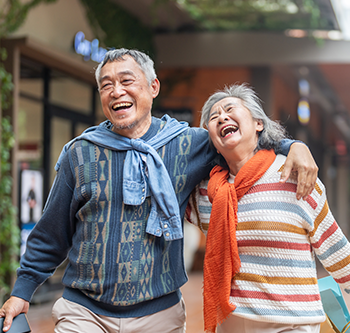Two older adults walk side by side along a pedestrian shopping street. One adult has an arm around the other’s shoulders. One carries a smartphone, and the other carries shopping bags. Storefronts, greenery, and out-of-focus buildings line the walkway. The image uses a shallow depth of field, with the people in focus and the background blurred.