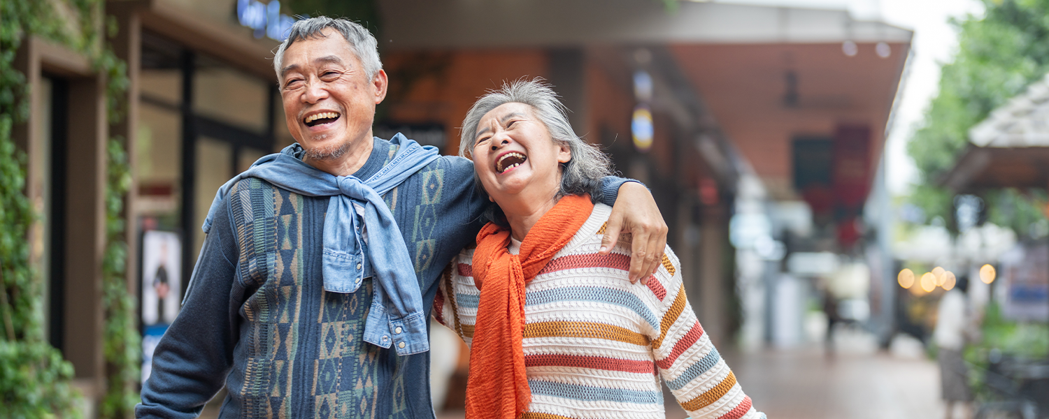 Two older adults walk side by side along a pedestrian shopping street. One adult has an arm around the other’s shoulders. One carries a smartphone, and the other carries shopping bags. Storefronts, greenery, and out-of-focus buildings line the walkway. The image uses a shallow depth of field, with the people in focus and the background blurred.