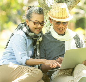 An elder couple sitting together against a tree in the park looking at their laptop