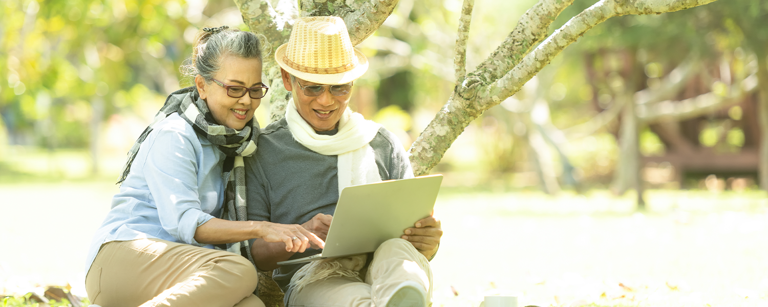 An elder couple sitting together against a tree in the park looking at their laptop