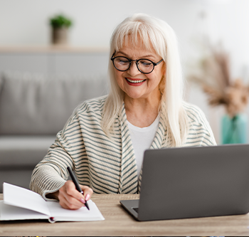 Older woman in a striped sweater smiling while writing in her journal with her laptop open