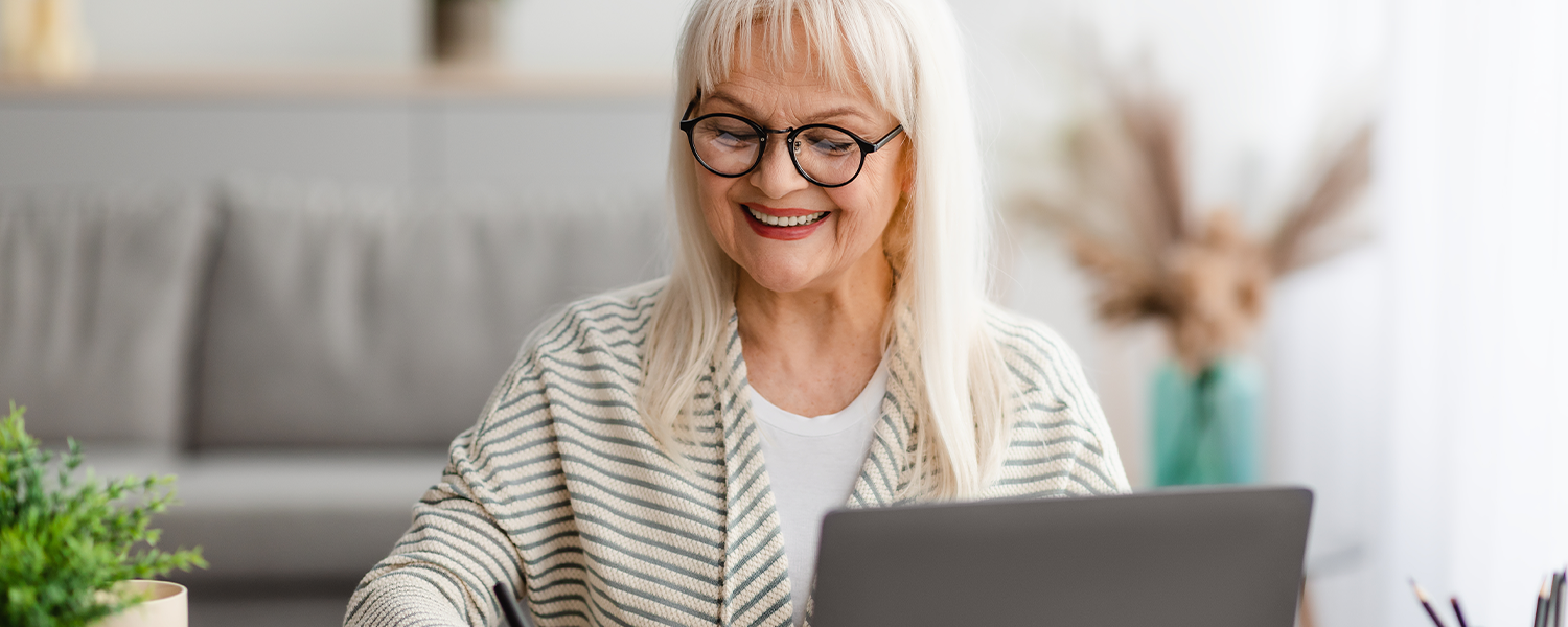 Older woman in a striped sweater smiling while writing in her journal with her laptop open
