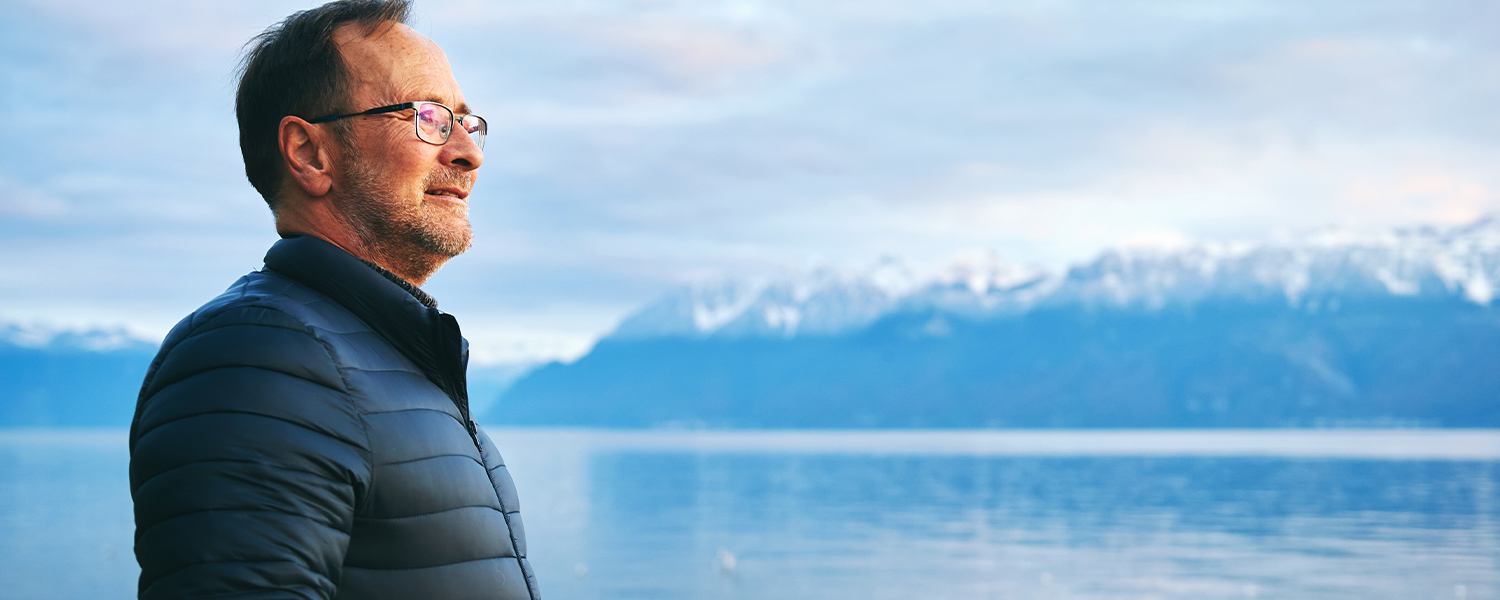 Man standing near a lake with mountains behind him looking off into the distance