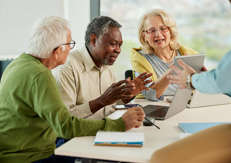 Adults seated around a table during a technology training session, using laptops, tablets, and smartphones while an instructor presents information on a tablet in a bright meeting room.