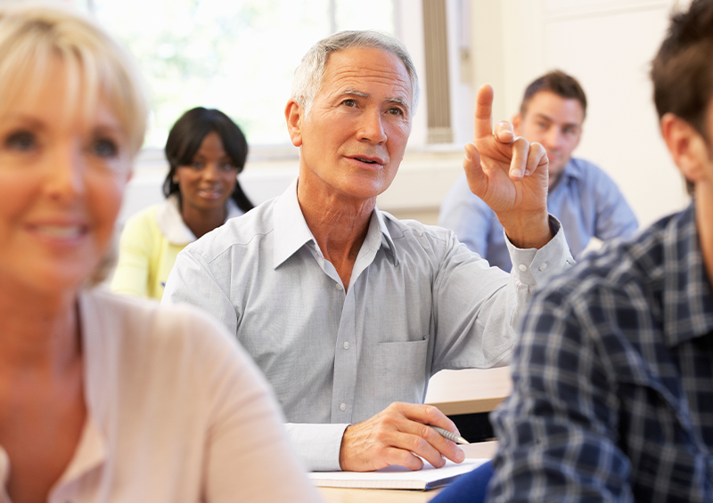 Participant in a classroom setting raises a hand to ask a question or contribute to a discussion, while other attendees sit in the background taking part in the session.