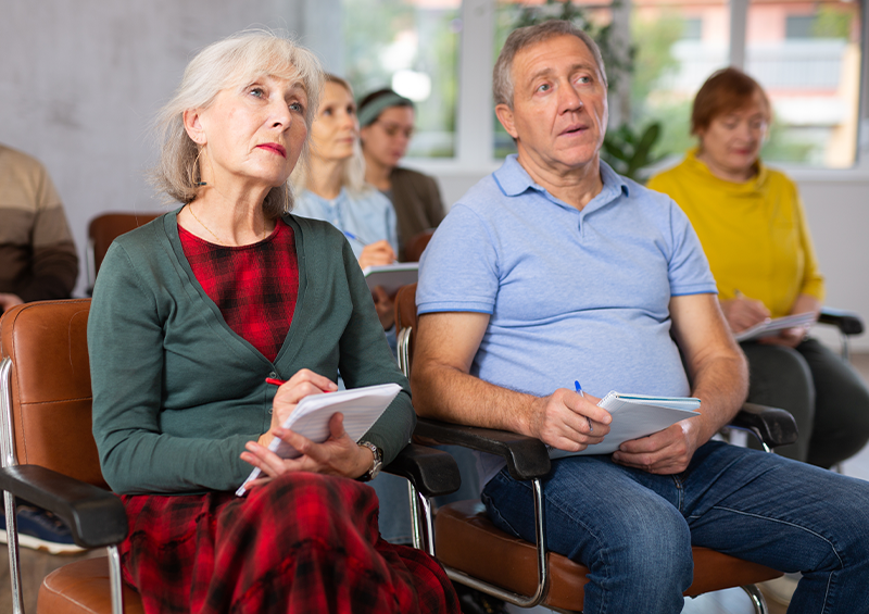 Group of adults seated in a training workshop, holding notebooks and taking notes while listening to a presentation in a classroom environment.