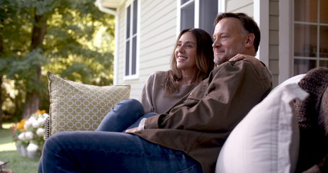 Alt text: Two people sit together on a front porch with cushioned outdoor seating, surrounded by a landscaped yard and the exterior of a light-colored home.
