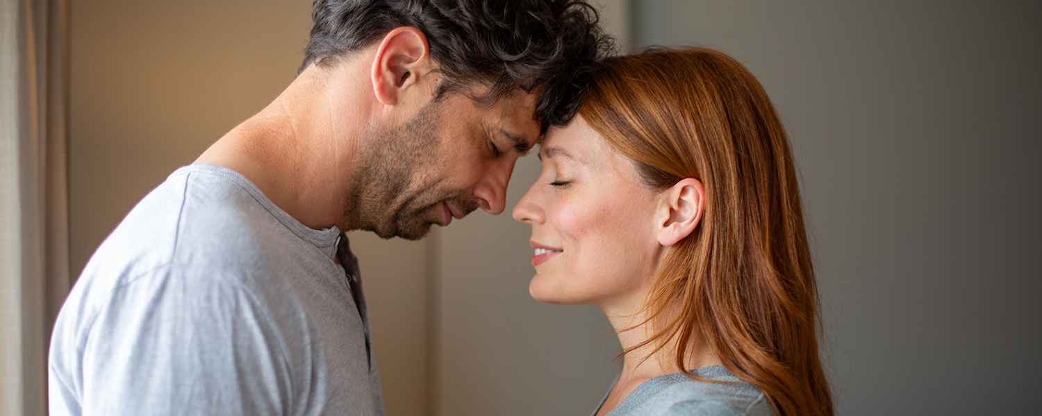 Two people standing close together indoors with their foreheads touching. One person with short dark curly hair wears a light gray shirt, and the other person with long reddish-brown hair wears a light blue top. The scene is softly lit with a blurred neutral-toned background.